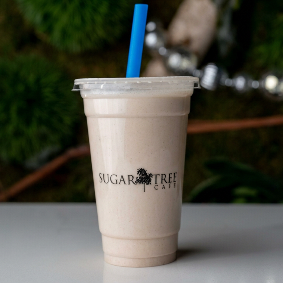 Sugar Tree Cafe smoothie in a clear cup with a blue straw, displayed on a table against leafy greenery.